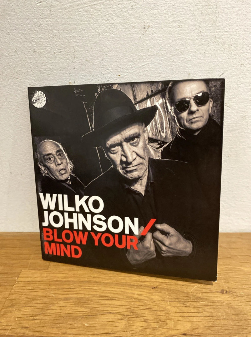 CD Album cover for Wilko Johnson's 'Blow Your Mind' on a wooden surface with a white wall background.
